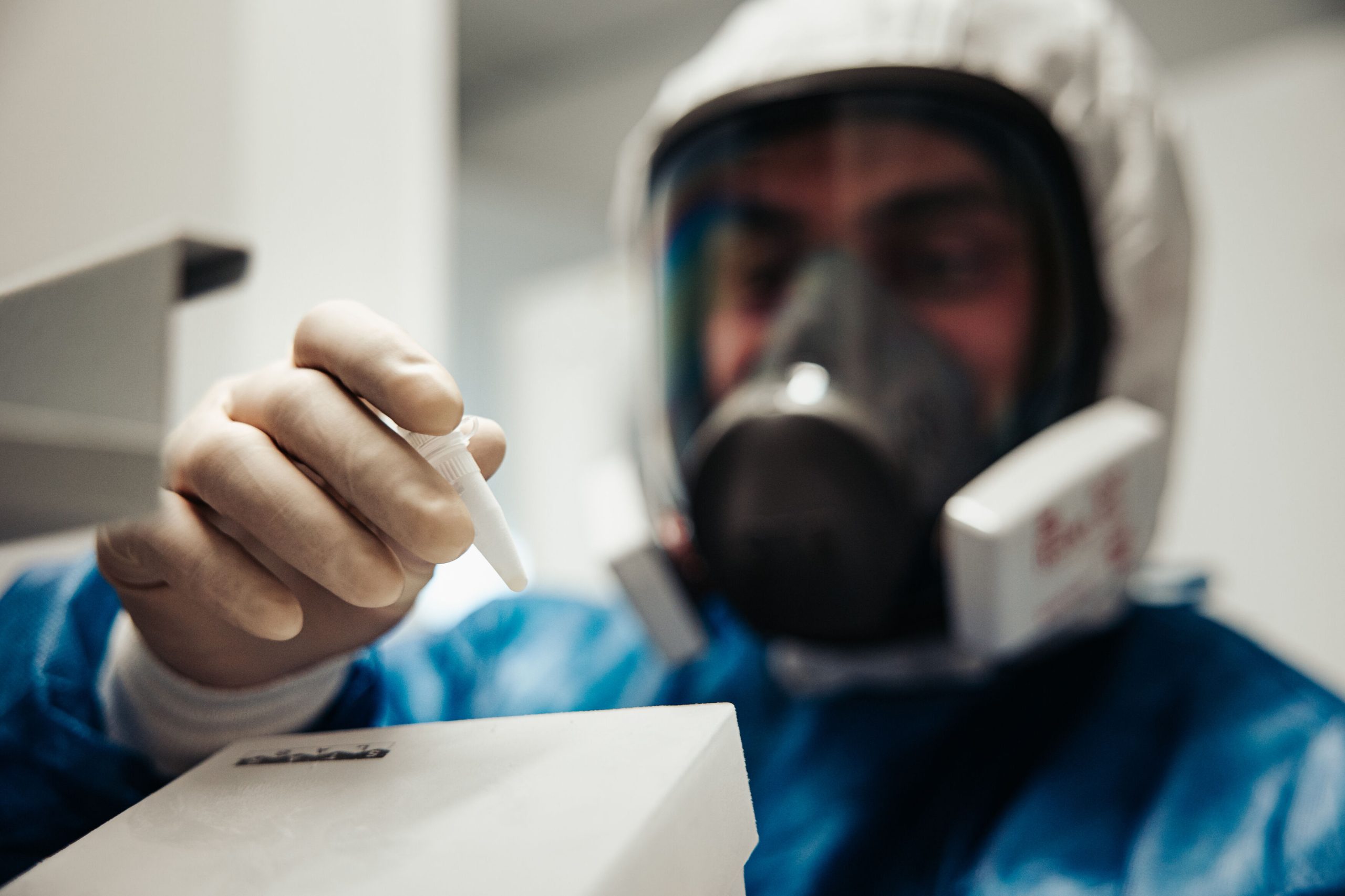 A researcher wearing a full face mask, with cap, gown and gloves, handling small sample vials.