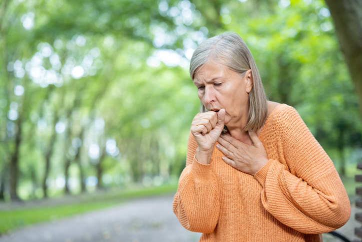 Photograph of a woman coughing outdoors in a park while holding their chest.