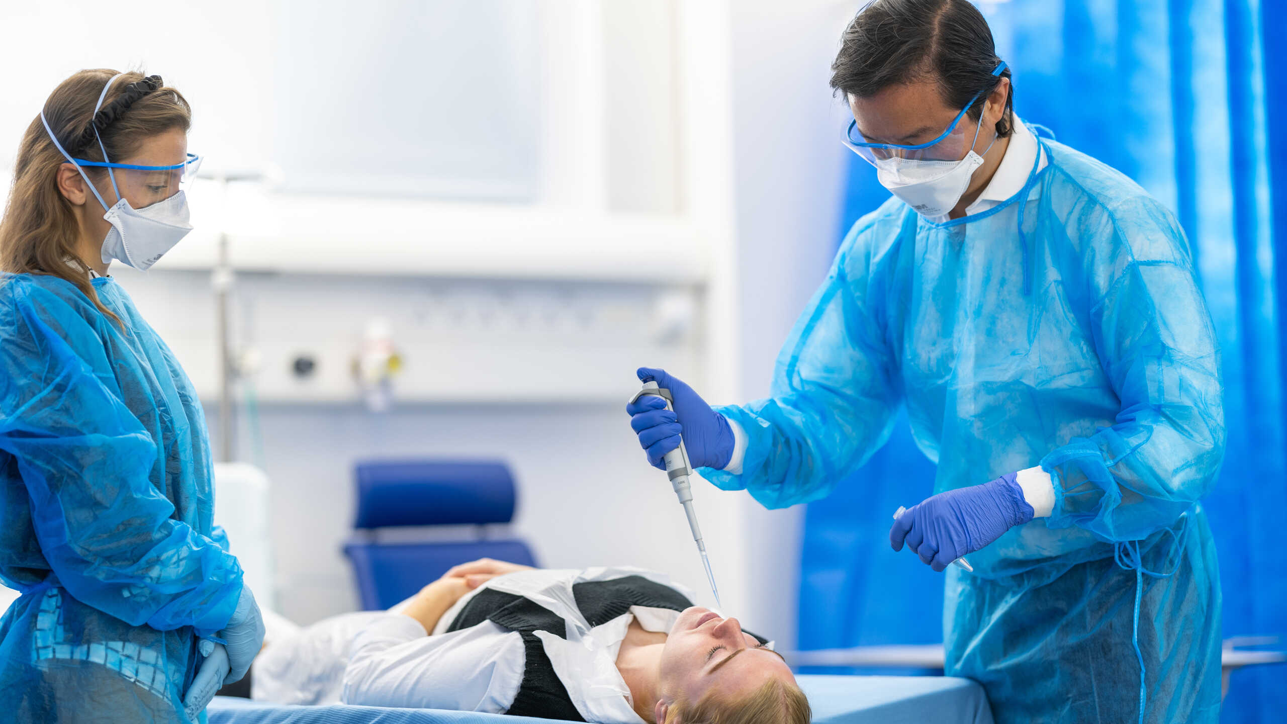 Photograph of two researchers in protective equipment stood either side of a patient laying down. One researcher is lowering a pipette to the patient's nose.