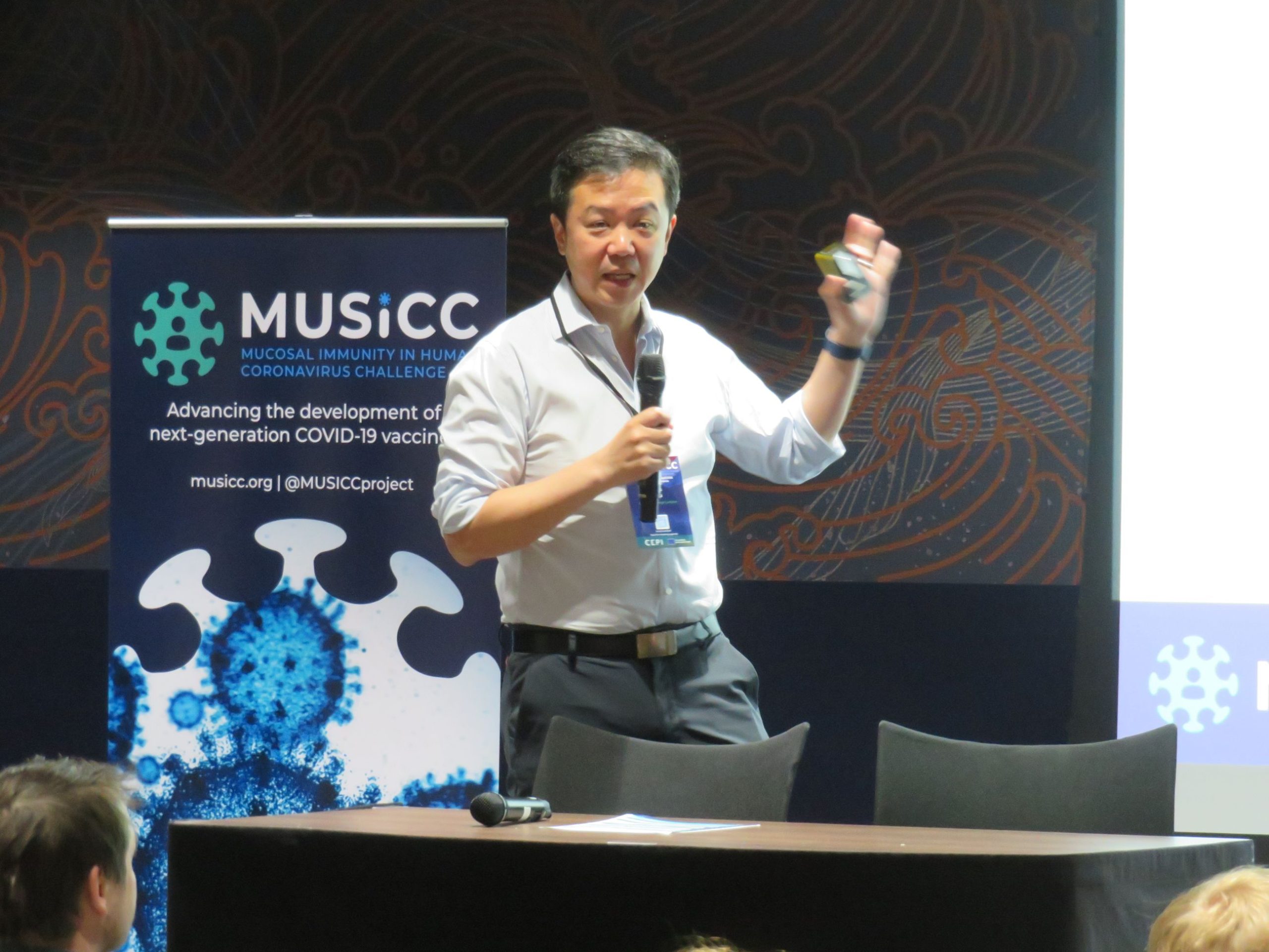 Photograph of Prof Chris Chiu presenting on stage at the 2026 Annual Meeting, with a MUSICC project banner in the background.