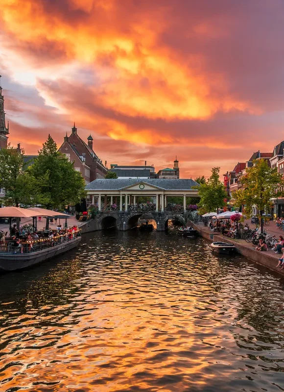 Photograph of sunset at city centre river. A bridge spans the river, with buildings either side and people sat on the banks.