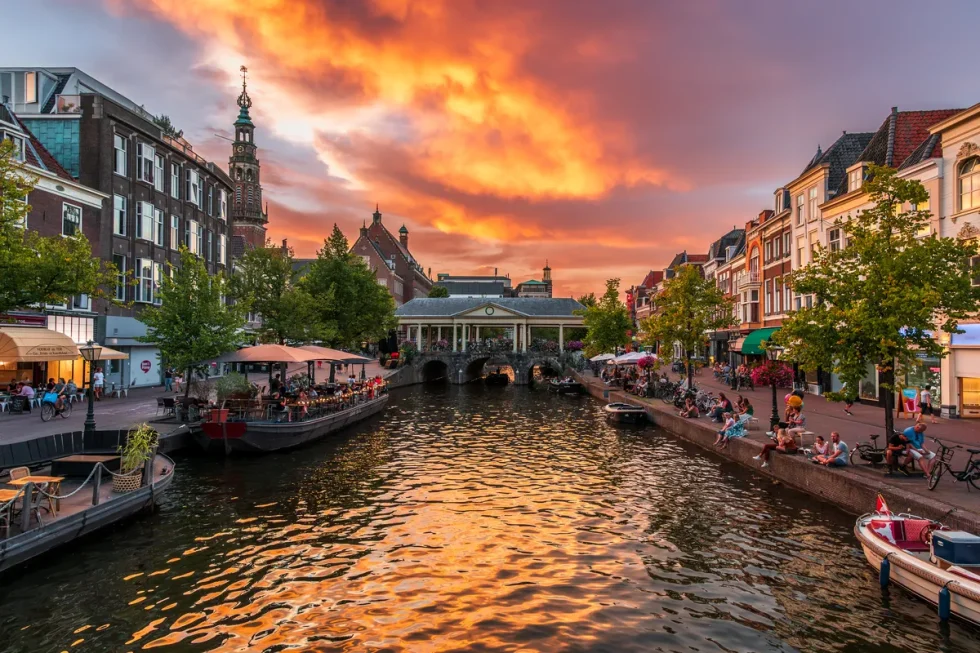 Photograph of sunset at city centre river. A bridge spans the river, with buildings either side and people sat on the banks.