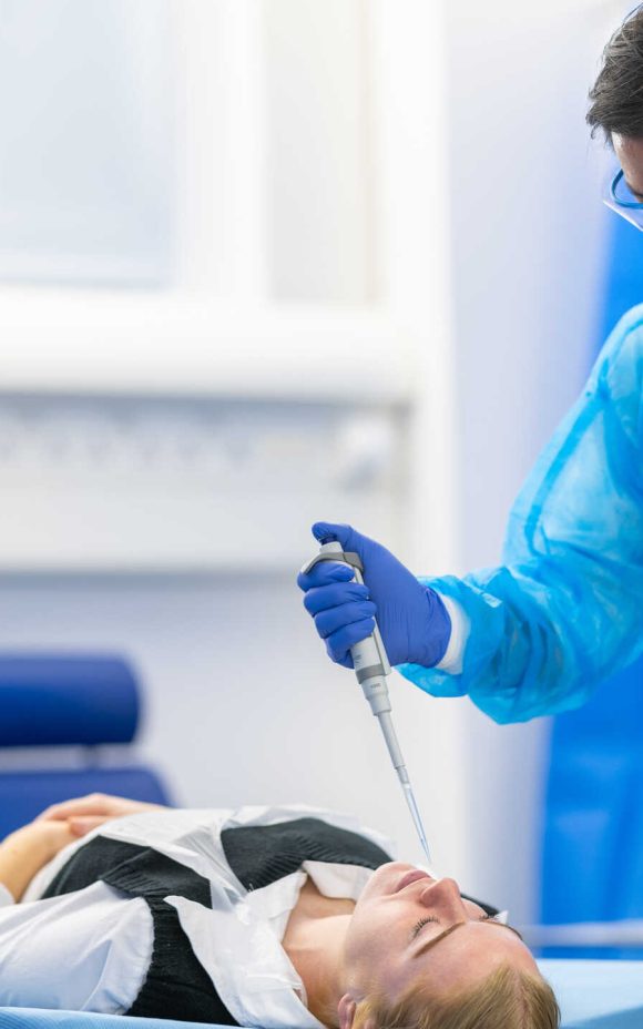 Photograph of two researchers in protective equipment stood either side of a patient laying down. One researcher is lowering a pipette to the patient's nose.
