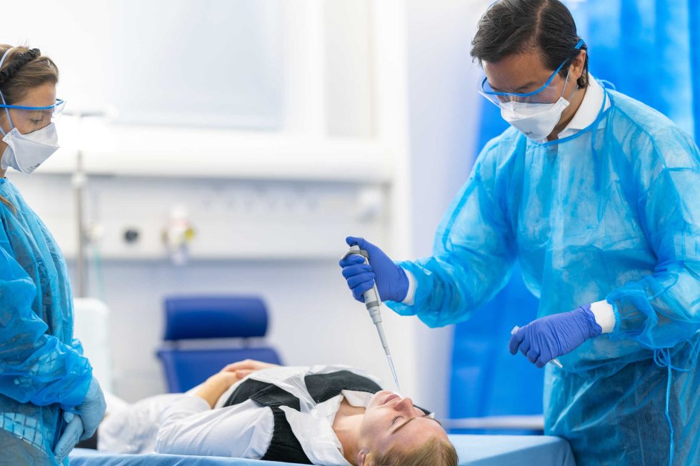Photograph of two researchers in protective equipment stood either side of a patient laying down. One researcher is lowering a pipette to the patient's nose.