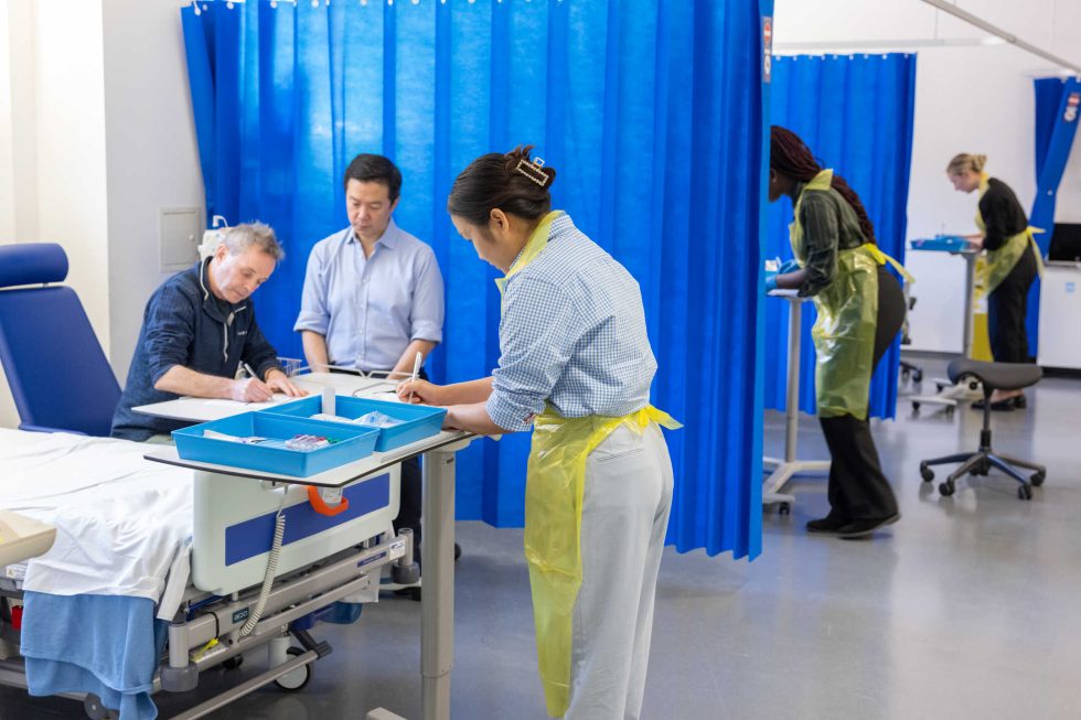 Photograph of a human challenge study. Multiple medical staff are monitoring patients and recording observations.